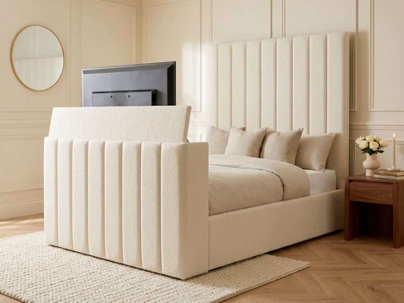 A photorealistic interior photograph of the Vertice TV Bed, a UK-made upholstered TV bed shown from a three-quarter rear/side angle so both the tall floor-standing fluted-channel headboard and matching footboard, dead straight vertical ribs, no curves and the foot end are visible, upholstered in Boucle fabric in a Cream colourway, dressed with a stone/cream duvet and three coordinating scatter cushions, set in a warm, softly lit bedroom with cream wainscoted panel-moulding walls, a round or octagonal gold-framed mirror on the left wall, a light oak/herringbone wood floor partly covered by a chunky cream jute or wool rug, a rich walnut wood nightstand to the right holding a small cream ceramic vase of white roses, a stack of two hardback books and a lit taper candle, soft natural daylight falling from an unseen window at a 45-degree angle, shallow depth of field, photorealistic interior-design magazine photography, shot on a full-frame camera at f/4, no people, no text, no watermark, no logos. The headboard and footboard must be entirely straight-edged and square-cornered &mdash; absolutely no curved, rounded, wave, or scalloped lines anywhere on the frame. Image size exactly 1500 x 1066 pixels, landscape orientation.